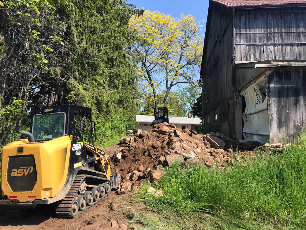 Excavator and track loader removing stones and dirt from an old barn foundation