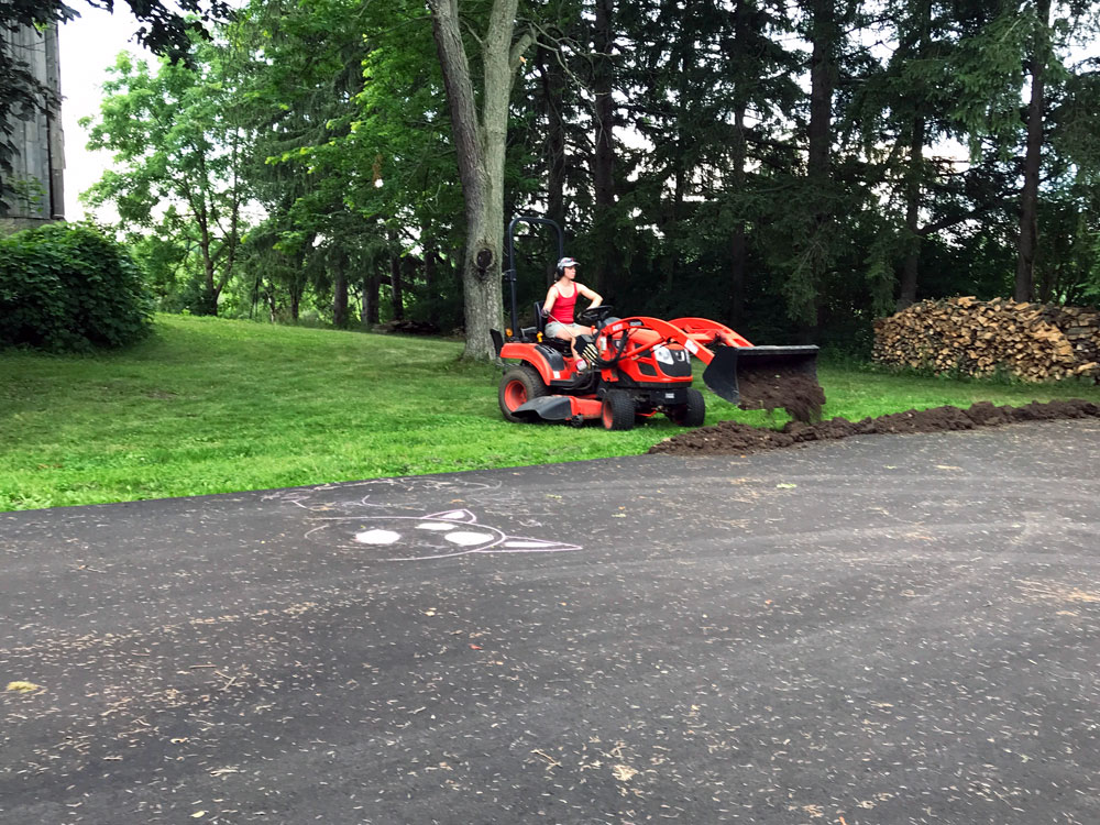 Woman on a small orange tractor dumping dirt alongside an asphalt driveway